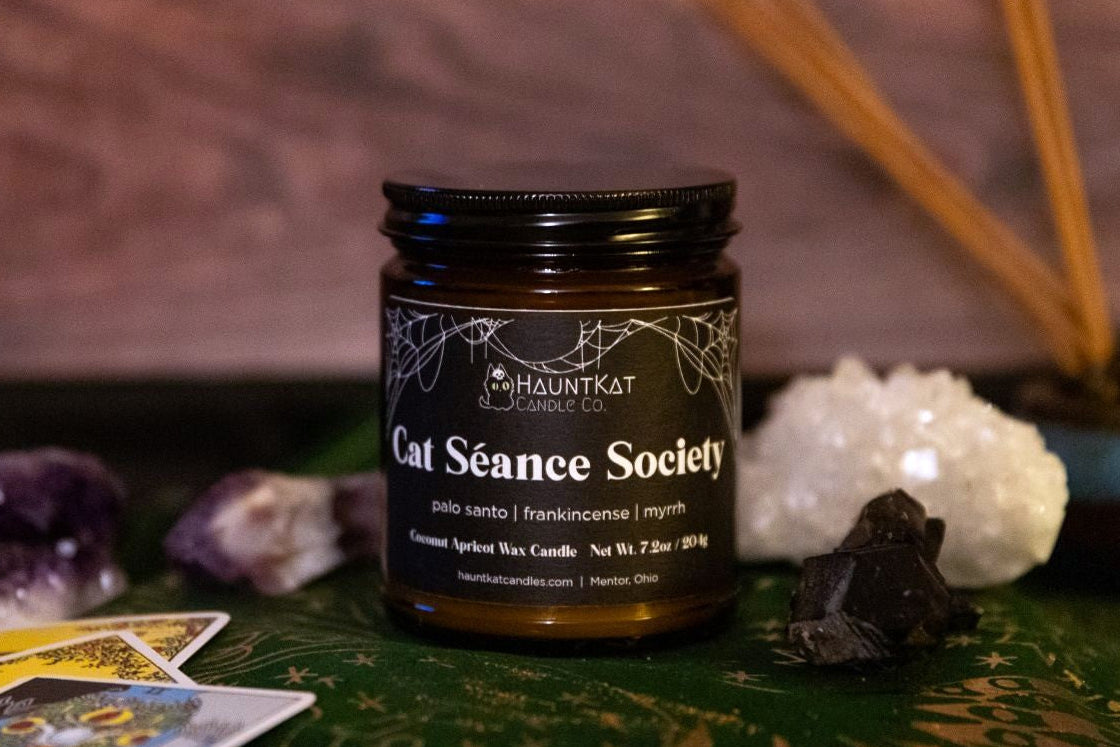 Candle jar labeled 'Cat Seance Society' on a mystical table with crystals and tarot cards.
