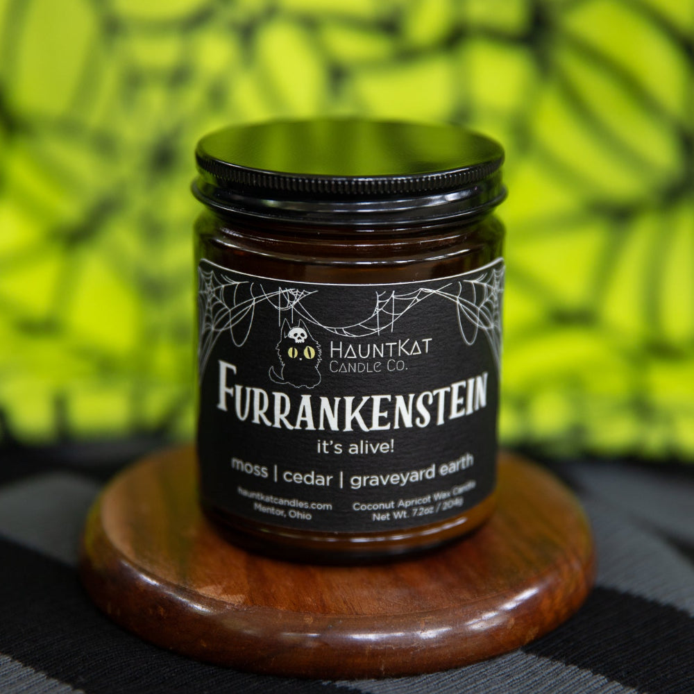 Amber candle named 'Furrankenstein' from Hauntkat Candle Co. on a wooden coaster with a green spiderweb background.