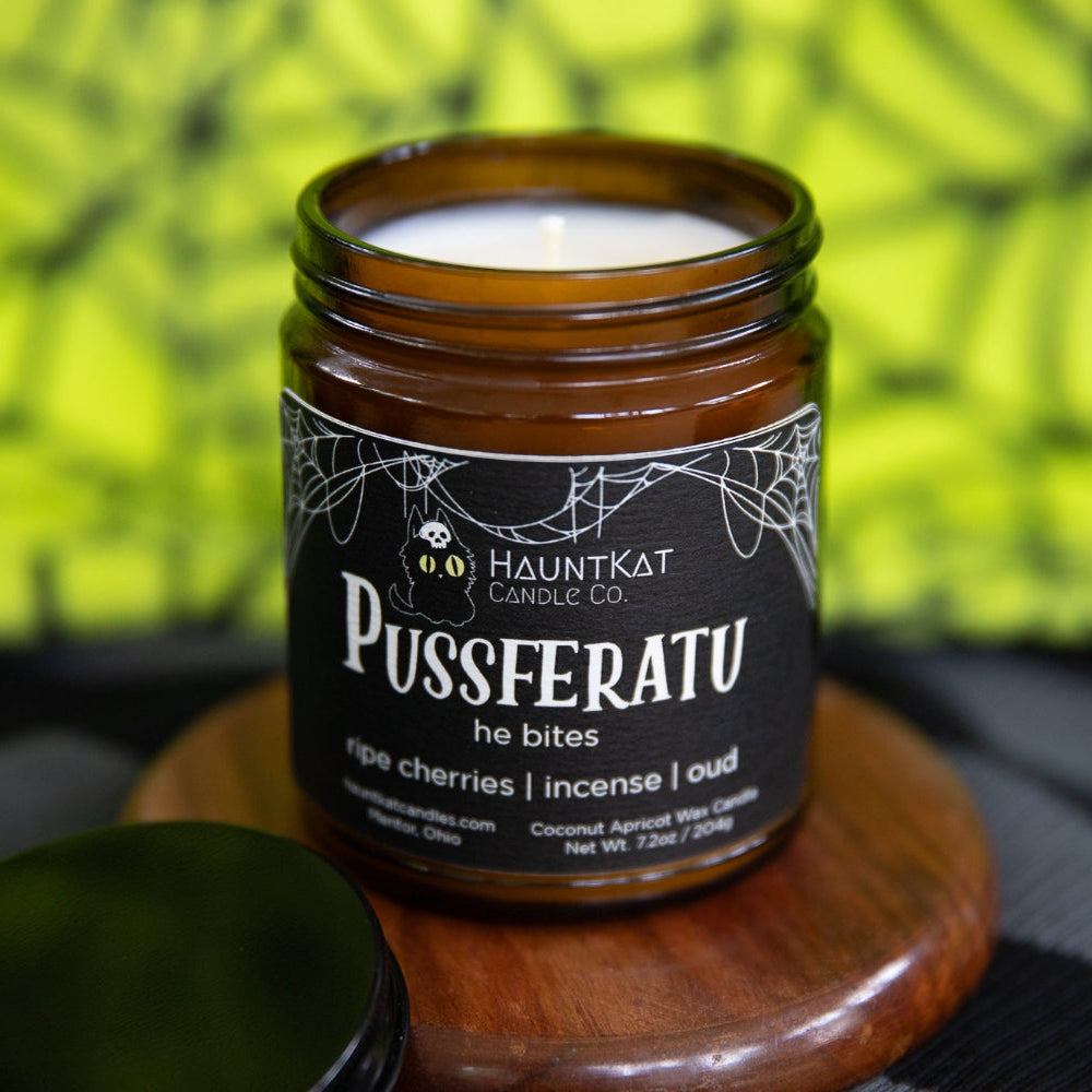 Candle jar labeled 'Pussferatu' by Hauntkat Candle Co. on a wooden coaster with a blurred green and black spiderweb background.
