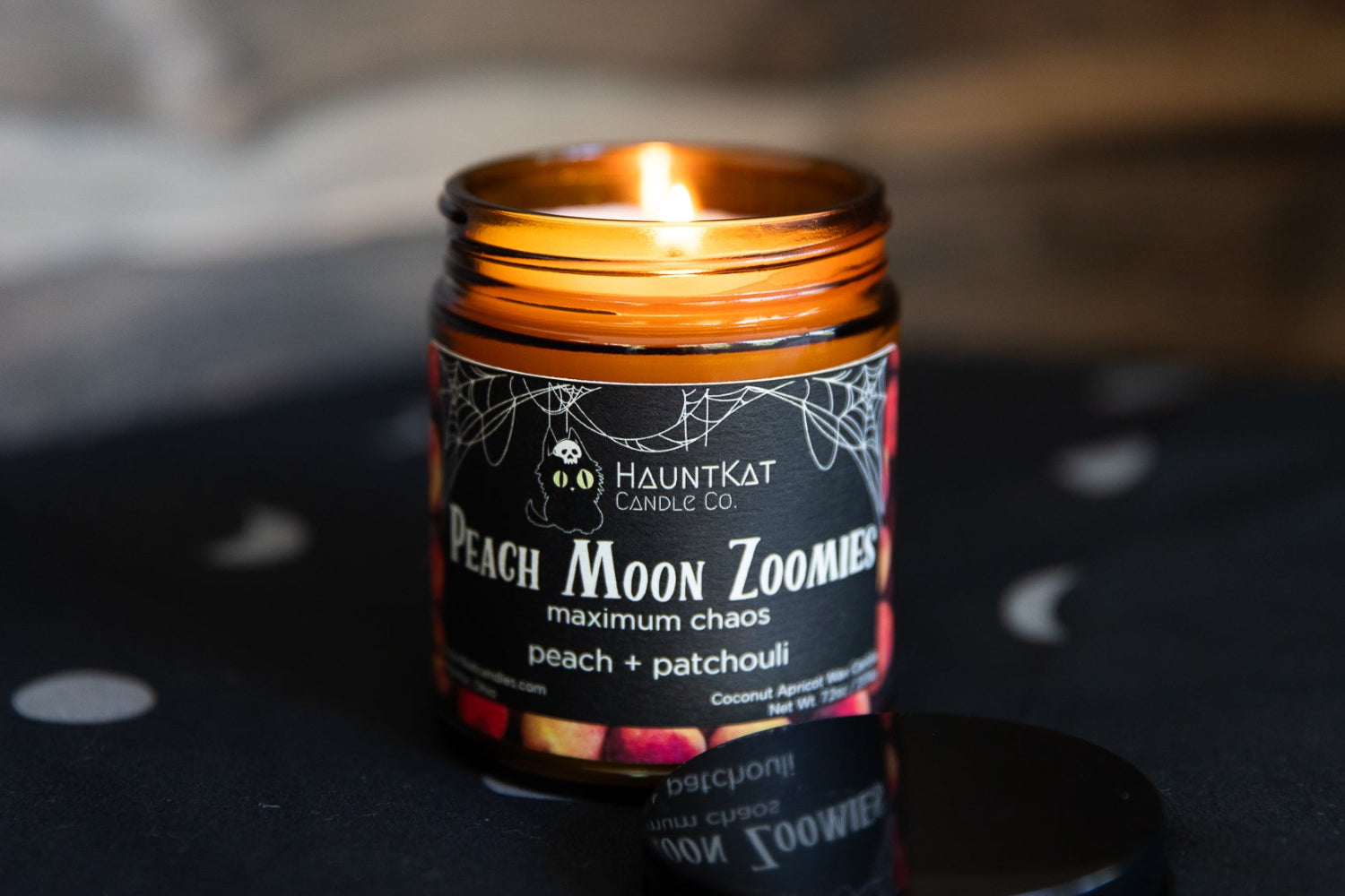 A lit amber glass jar candle with a black label reading 'Haunted Kat Candle Co. Peach Moon Zoomies maximum chaos peach + patchouli,' shown on a dark surface with a matching lid in front.