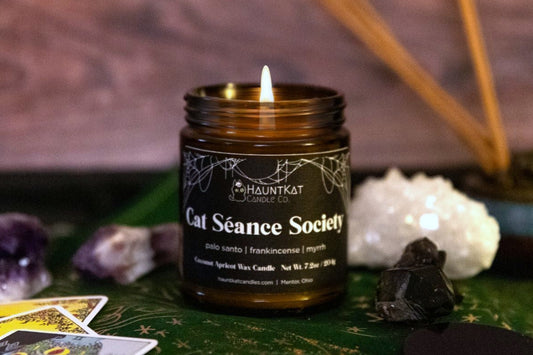 Candle in an amber jar with black label reading 'Cat Séance Society' on a mystical setting with tarot cards, crystals, and incense.