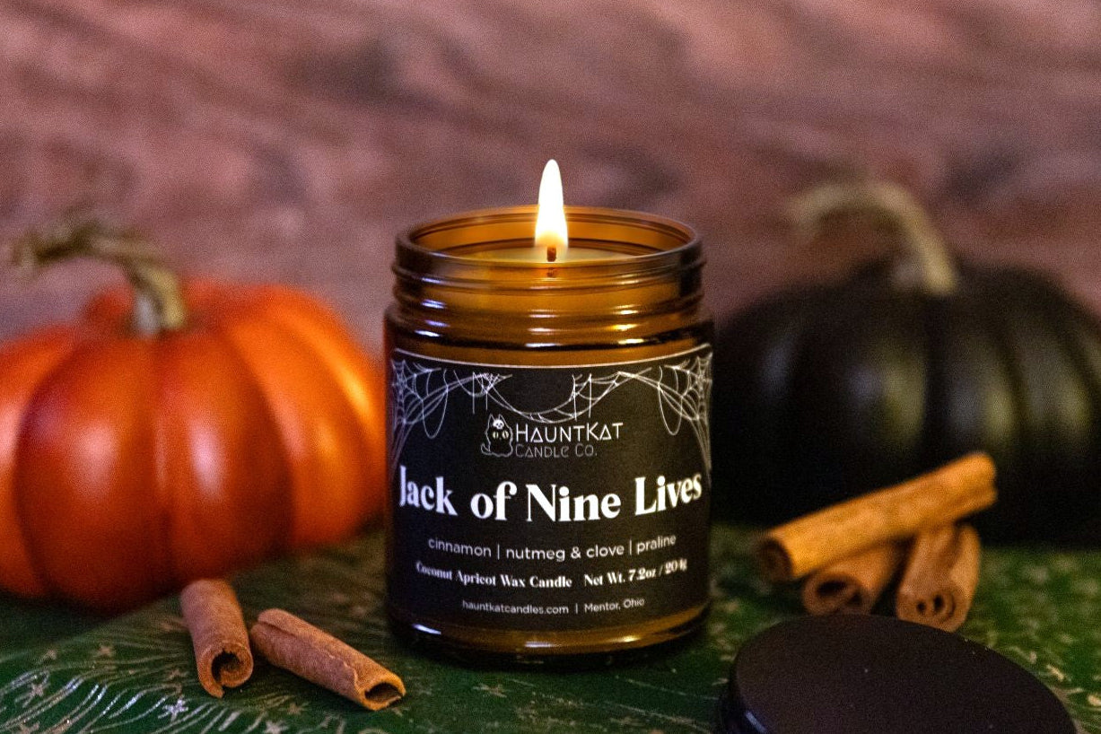 Candle in a glass jar labeled 'Jack of Nine Lives' with pumpkins and cinnamon sticks on a wooden surface.