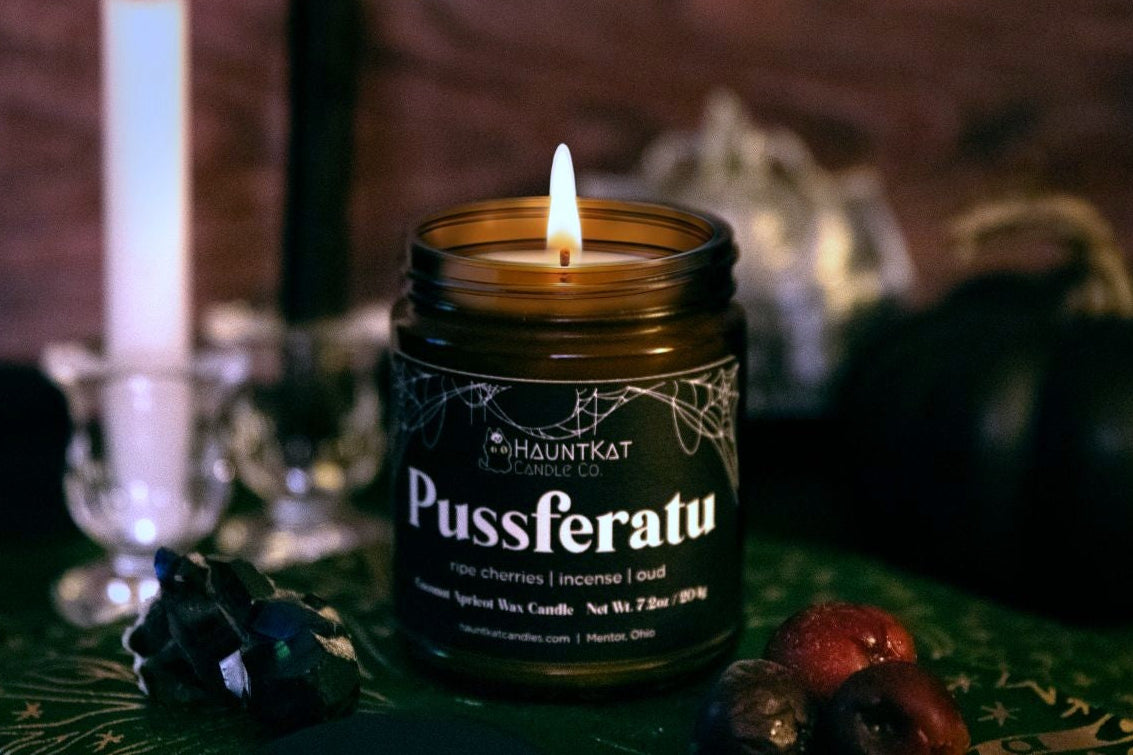Candle labeled 'Pussferatu' on a dark background with other candles and decor.
